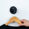 Hand die een houten kledinghanger met zwart kledingstuk vasthoudt, hangend aan een zwarte haak aan een witte muur