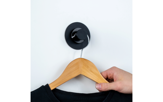 Hand die een houten kledinghanger met zwart kledingstuk vasthoudt, hangend aan een zwarte haak aan een witte muur