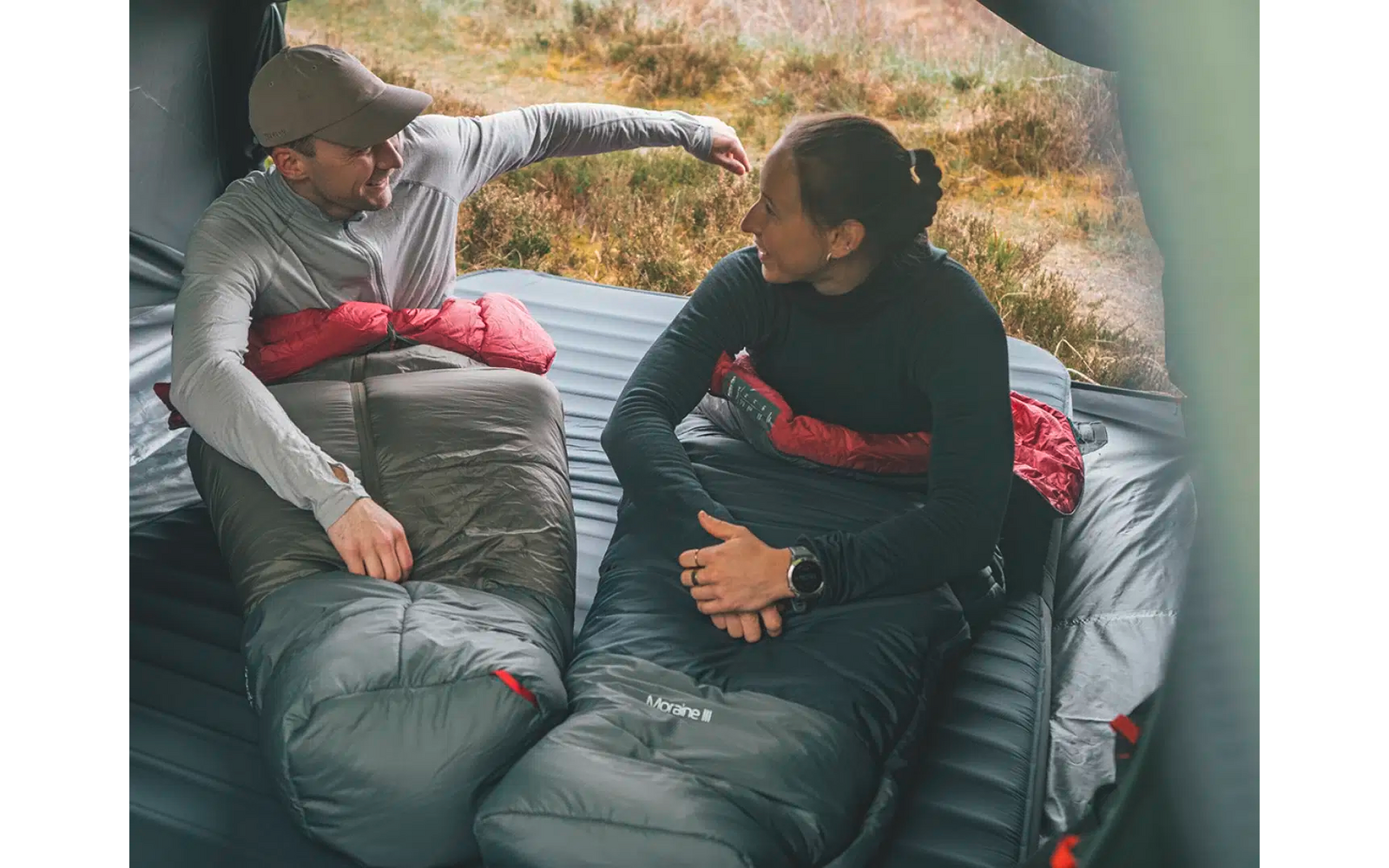 Twee personen zitten in slaapzakken op de grond, gras en stenen op de achtergrond