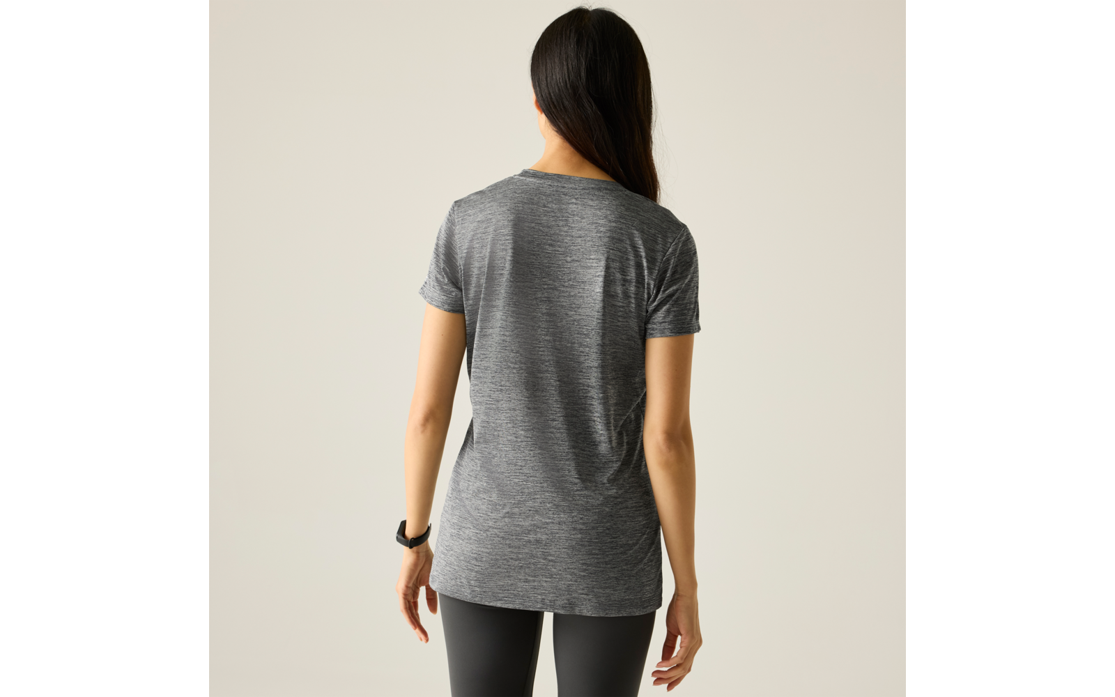 Personne de dos avec cheveux longs et foncés, t‑shirt gris à manches courtes, legging noir, poignet gauche avec bande noire, fond uni clair