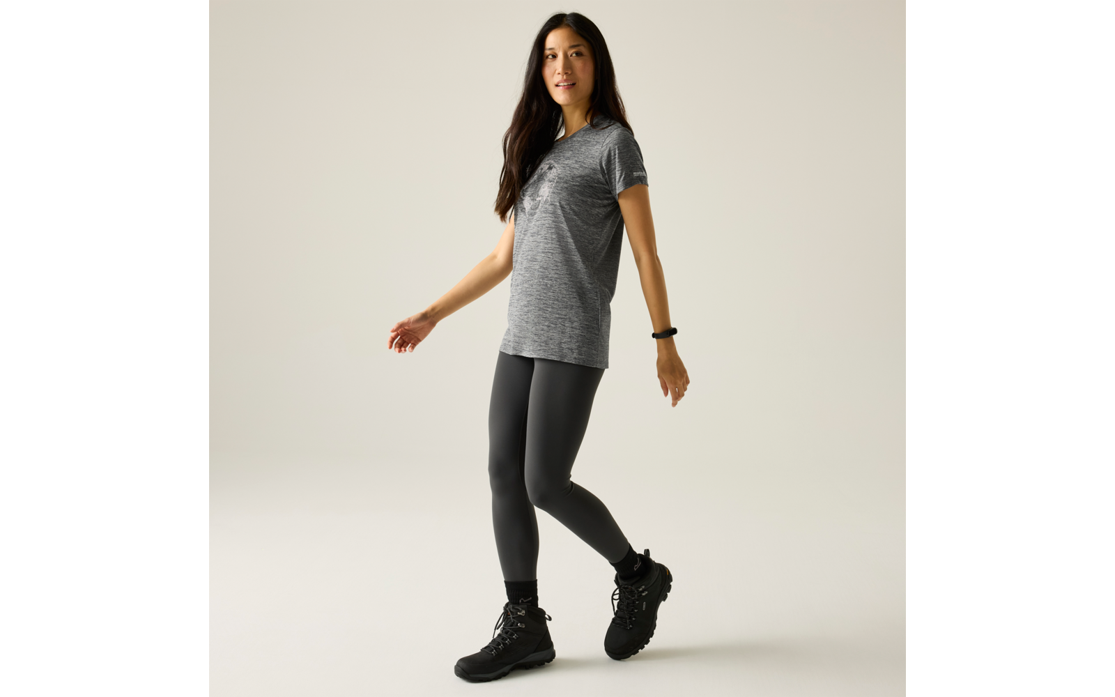 Femme aux longs cheveux foncés, portant un t-shirt gris à manches courtes, des leggings noirs et des chaussures de randonnée noires, debout un pied devant l'autre sur un fond uni clair; bracelet noir à un poignet