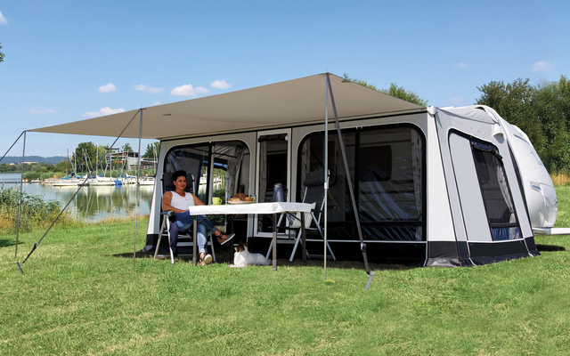 Caravane avec auvent sur une pelouse au bord d'un lac ; sous l'auvent une personne est assise à une table, un petit chien est couché sous la table ; en arrière-plan eau, bateaux et arbres