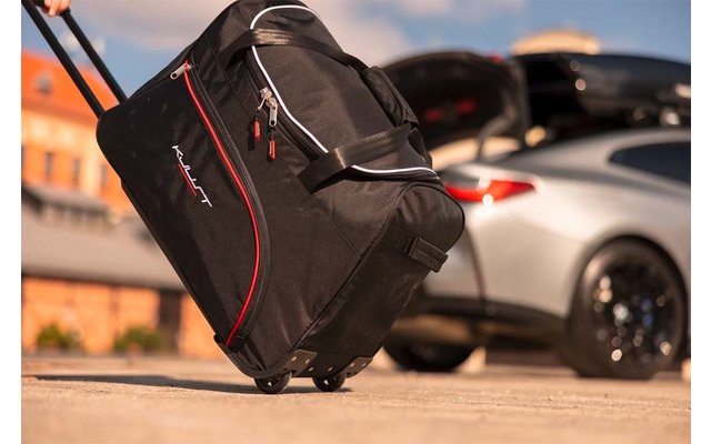 Sac de voyage noir à roulettes avec liseré rouge et poignée sortie sur sol pavé ; coffre ouvert d'une voiture de sport argentée en arrière-plan