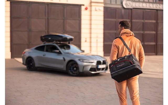 Personne de dos avec sac de voyage noir, marchant vers une voiture argentée stationnée avec coffre de toit devant des portes de garage fermées