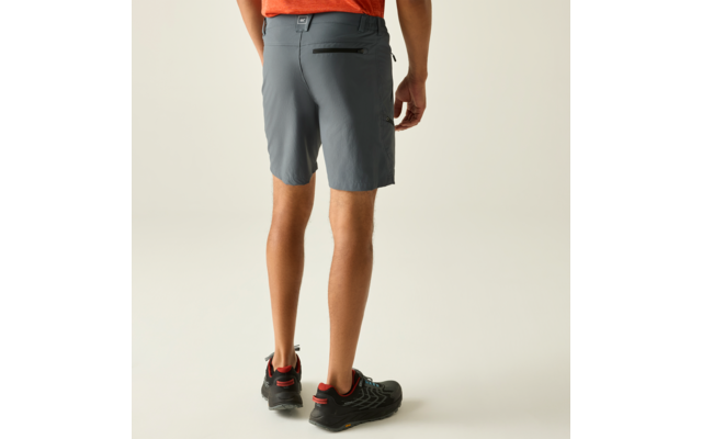 Rückansicht einer Person von der Taille abwärts in grauen Shorts mit Reißverschlusstasche hinten, teilweise sichtbares orangefarbenes Oberteil, schwarze Sportschuhe mit roten Akzenten, heller Hintergrund