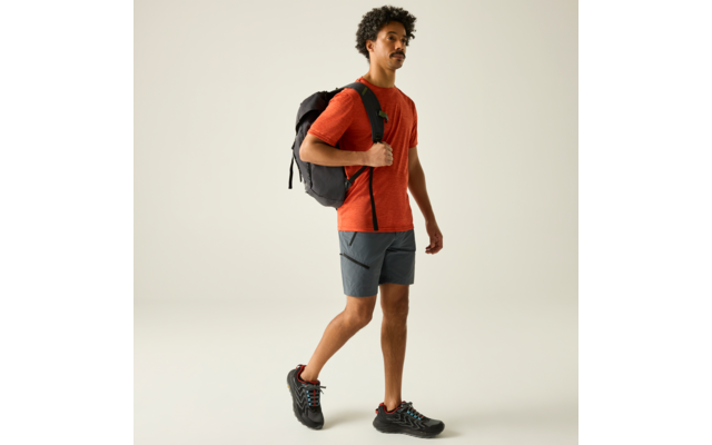 Mann mit Rucksack, rotes T‑Shirt, graue Shorts, schwarze Schuhe, geht vor hellem Hintergrund