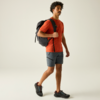 Mann mit Rucksack, rotes T‑Shirt, graue Shorts, schwarze Schuhe, geht vor hellem Hintergrund