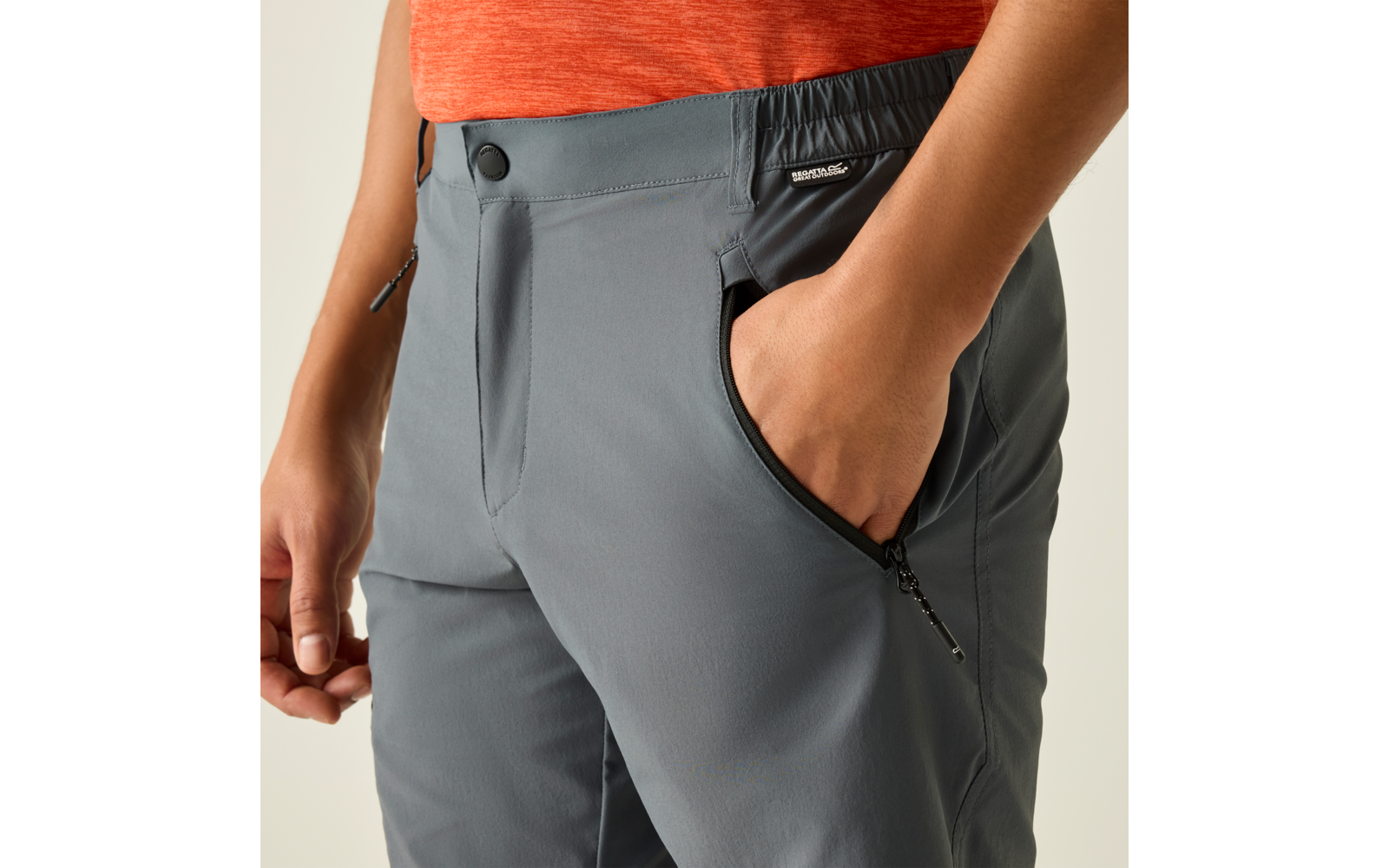 Oberkörper bis Oberschenkel einer Person in orangefarbenem T-Shirt und grauer Hose; rechte Hand in vorderer Reißverschlusstasche, linker Arm seitlich