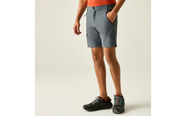 Stehende Person, unterer Bildausschnitt: graue Shorts, sichtbares orangefarbenes Oberteil, Hände in den Taschen, schwarze Wanderschuhe mit blauen und roten Schnürsenkeln, heller einfarbiger Hintergrund