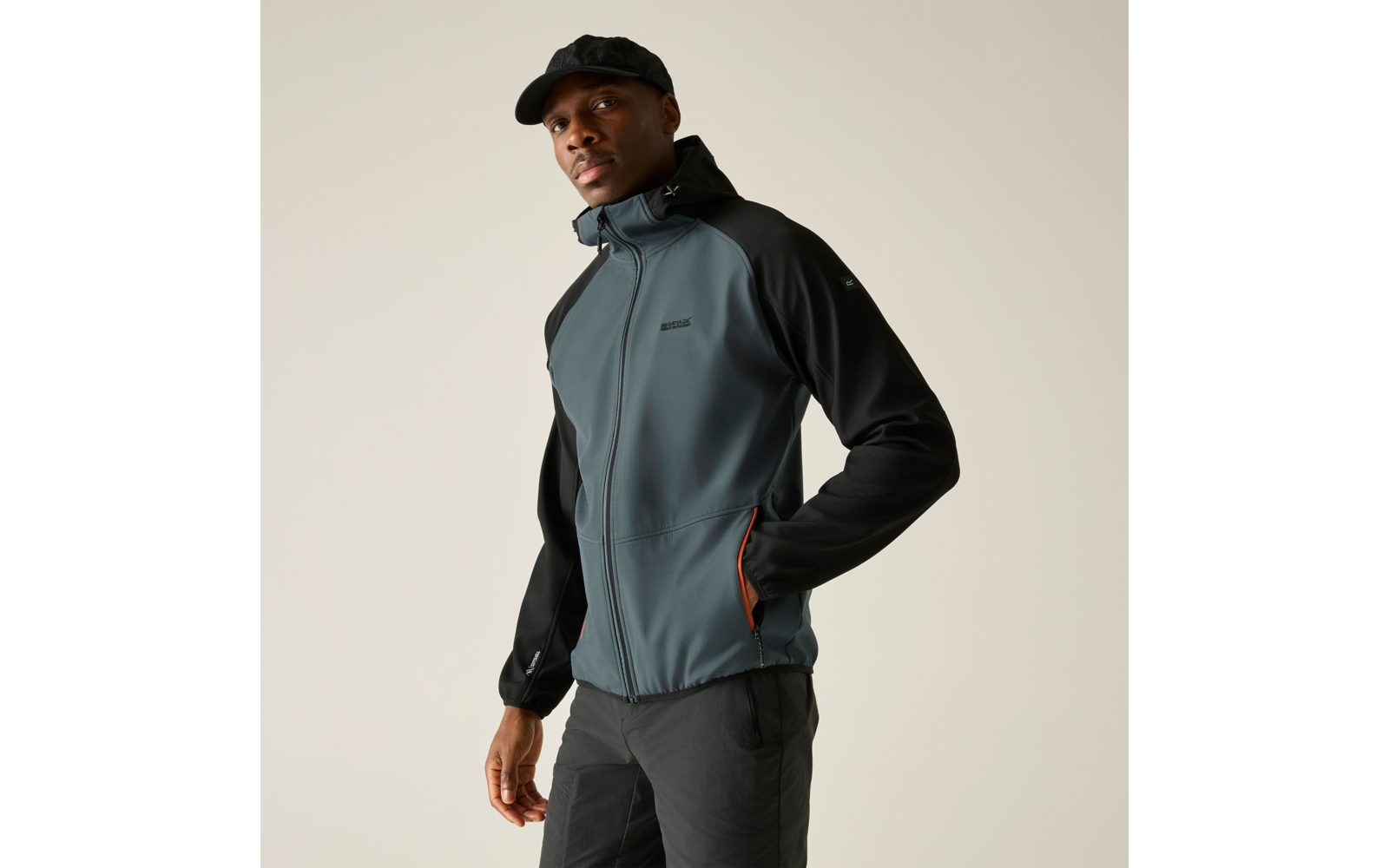 Person mit schwarzer Schirmmütze, grauschwarzer Reißverschlussjacke mit orangefarbenem Taschenfutter, eine Hand in der Tasche, vor hellem einfarbigem Hintergrund