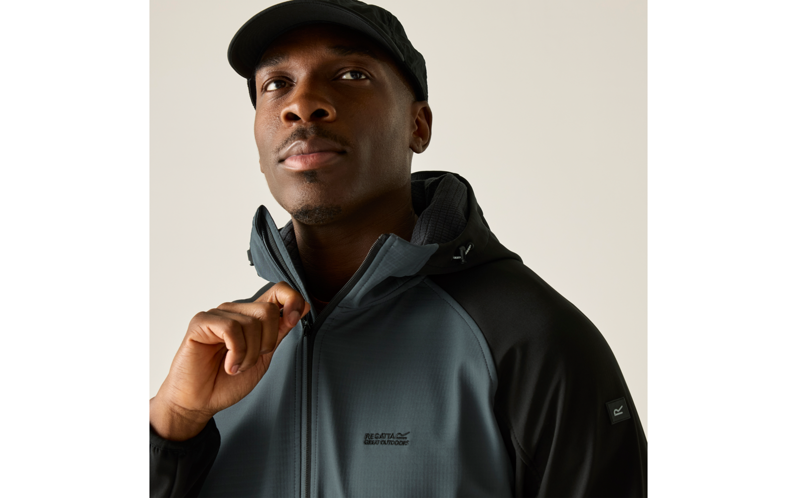 Person mit schwarzer Kappe und dunkelgrauer Kapuzenjacke zieht mit einer Hand den Reißverschluss hoch. Heller einfarbiger Hintergrund