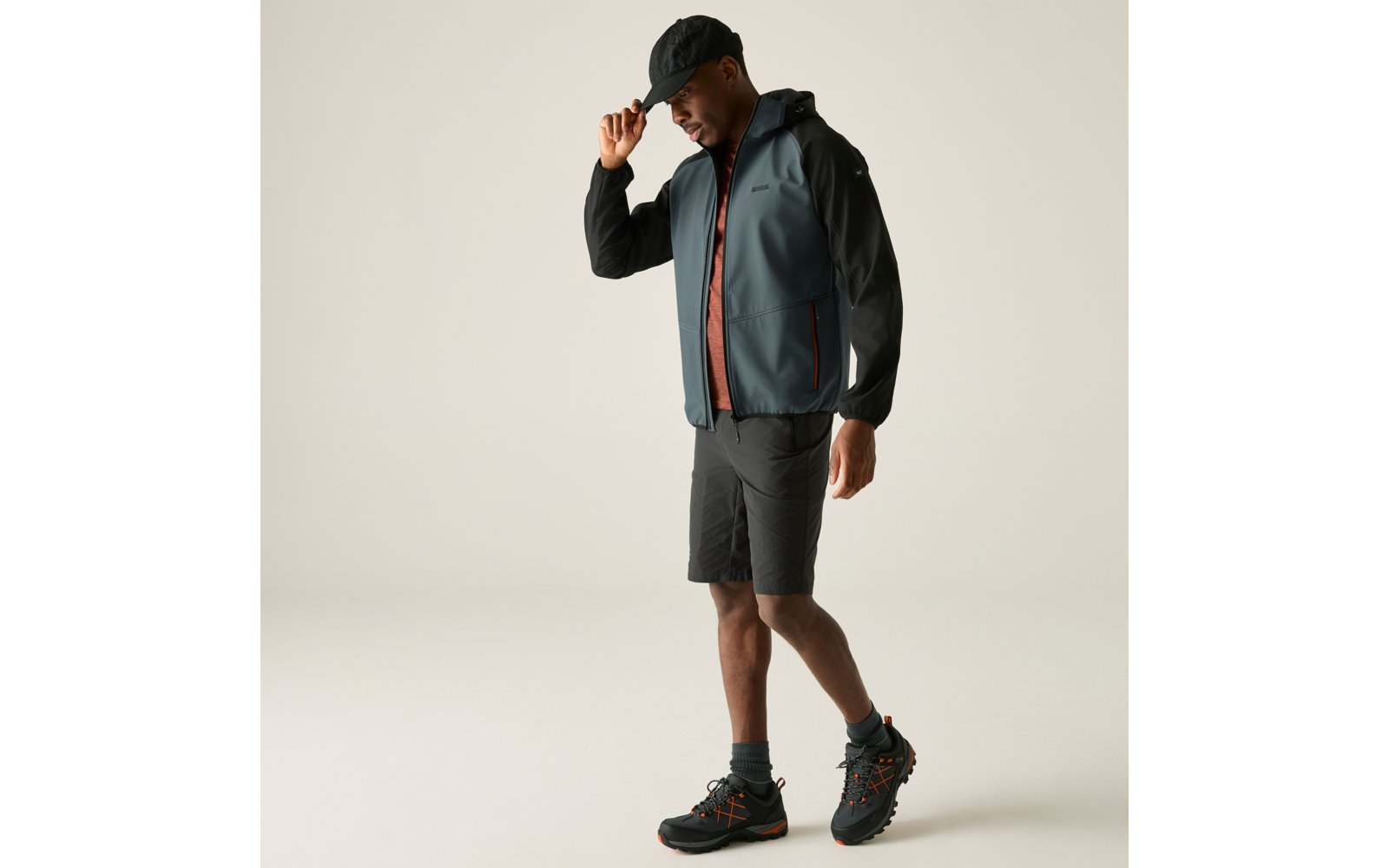 Eine stehende Person, die mit der Hand eine schwarze Kappe anfasst, trägt eine graue Jacke mit schwarzer Kapuze, ein rotes T-Shirt, schwarze Shorts und schwarze Wanderschuhe mit orangefarbenen Details