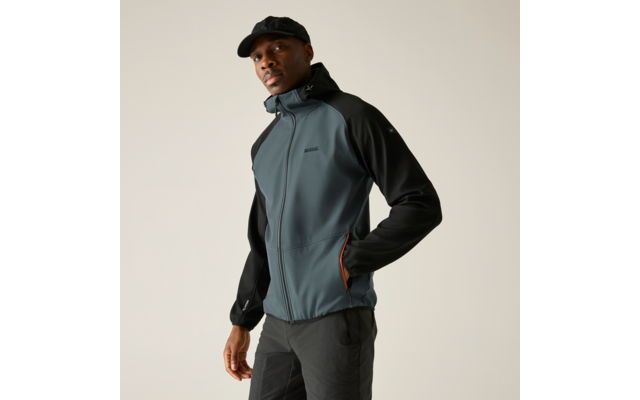 Person mit schwarzer Schirmmütze, grauschwarzer Reißverschlussjacke mit orangefarbenem Taschenfutter, eine Hand in der Tasche, vor hellem einfarbigem Hintergrund