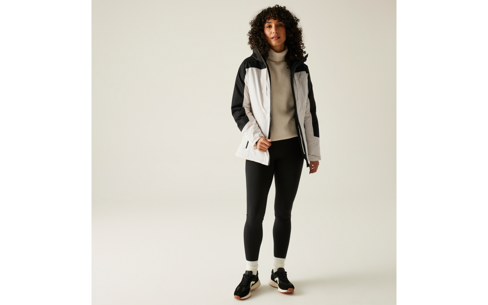 Vrouw met krullend haar draagt een wit-zwart sportjack, zwarte joggingbroek en sportschoenen, staat voor een lichte achtergrond