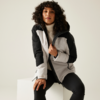Personne aux cheveux bouclés et foncés assise sur un cube blanc, portant une veste zippée gris clair et noire avec capuche, un pull à col roulé blanc et un pantalon noir, arrière-plan clair uni