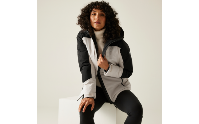 Personne aux cheveux bouclés et foncés assise sur un cube blanc, portant une veste zippée gris clair et noire avec capuche, un pull à col roulé blanc et un pantalon noir, arrière-plan clair uni