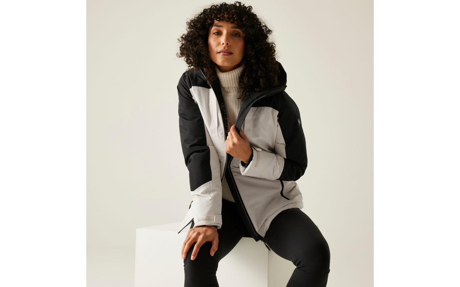 Personne aux cheveux bouclés et foncés assise sur un cube blanc, portant une veste zippée gris clair et noire avec capuche, un pull à col roulé blanc et un pantalon noir, arrière-plan clair uni