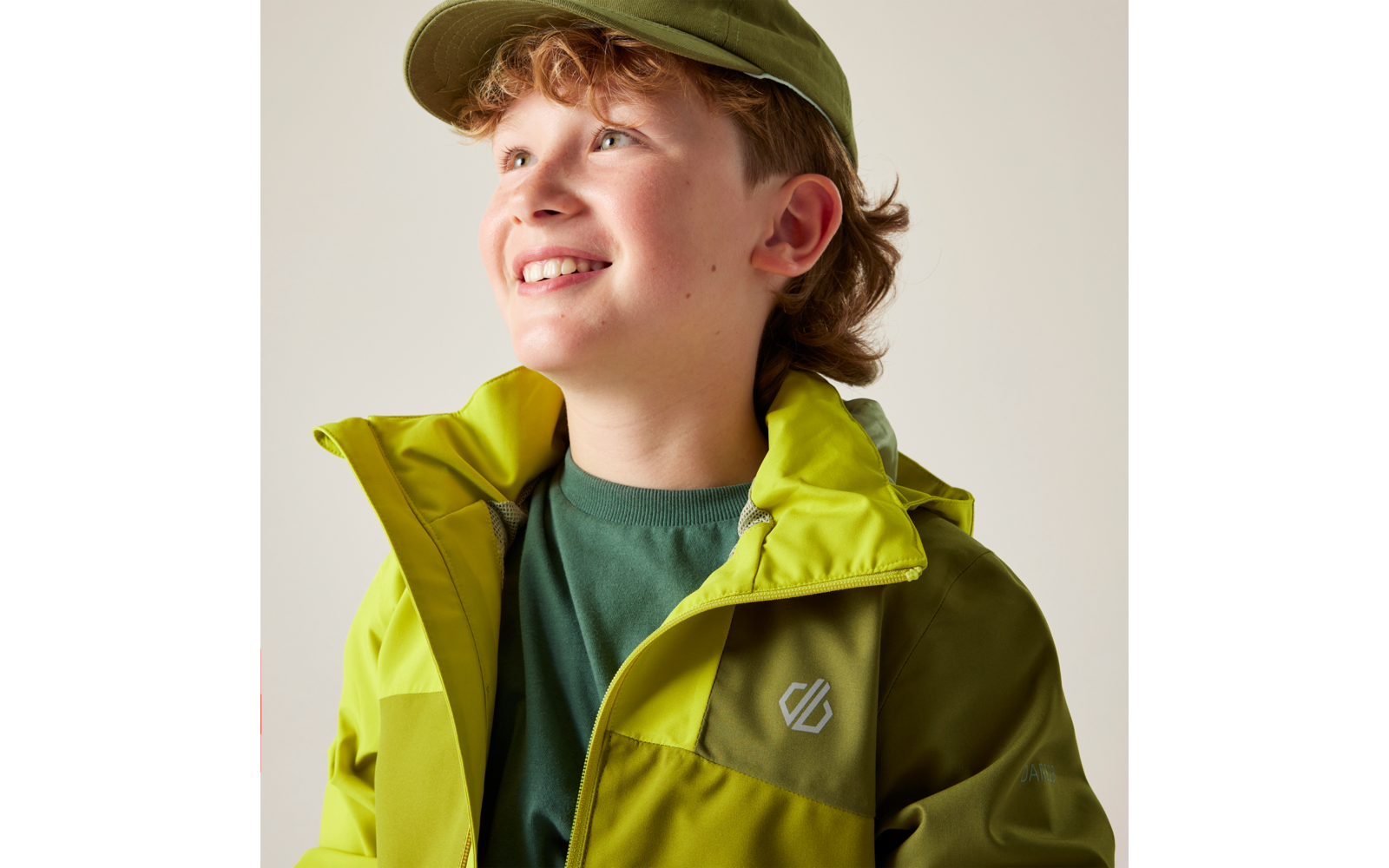 Person mit grüner Mütze und zweifarbiger grüner Jacke, mit einem Lächeln, Blick nach oben rechts, heller Hintergrund