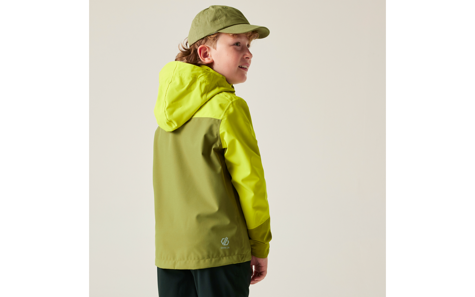 Junge von hinten, trägt zweifarbige grüne Jacke mit Kapuze und grüne Kappe, blickt über die Schulter vor hellem Hintergrund