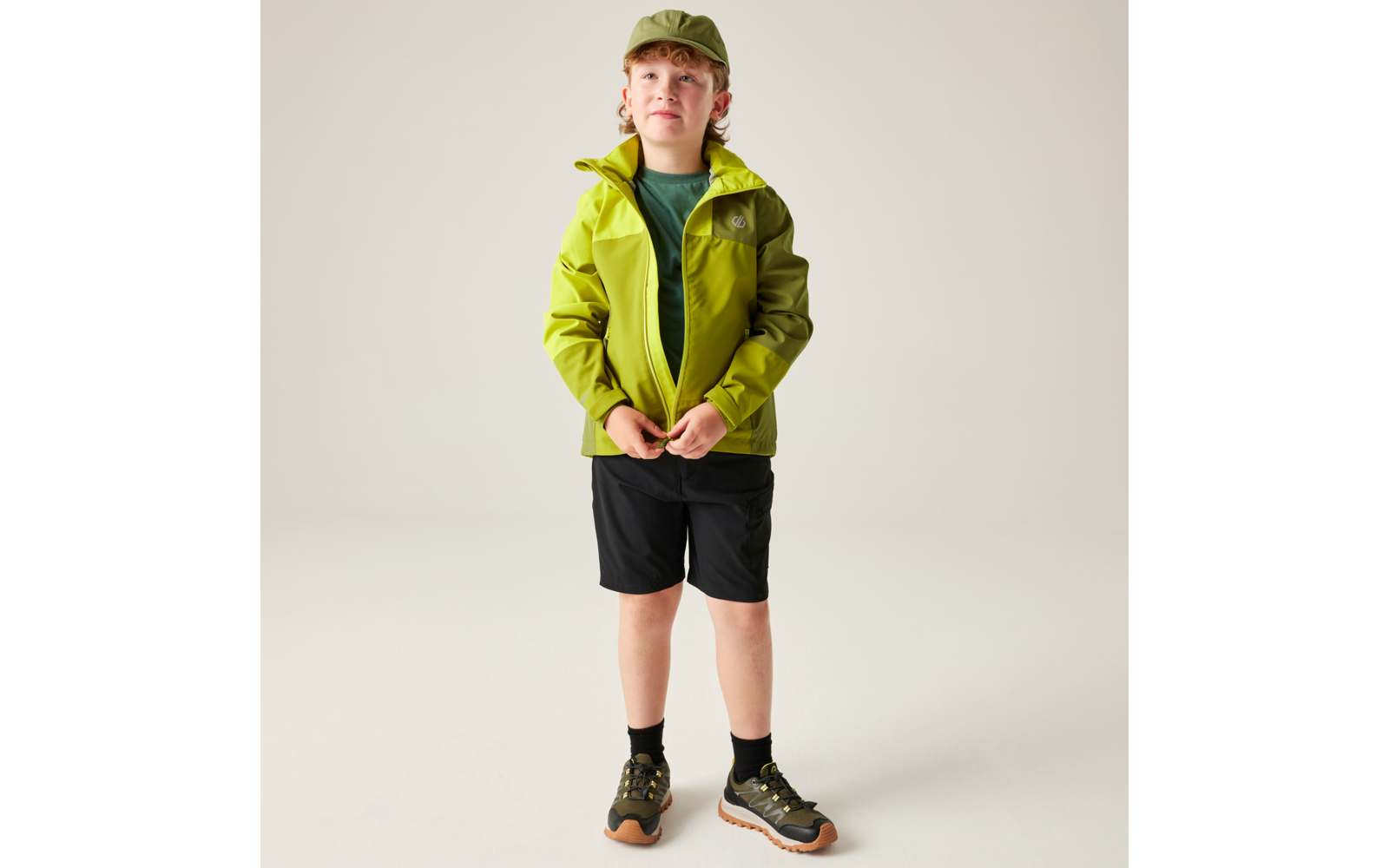 Kind steht, trägt olivgrüne Jacke und Kappe, dunkles T-Shirt, schwarze Shorts, schwarze Socken und grün-braune Schuhe vor hellem Hintergrund