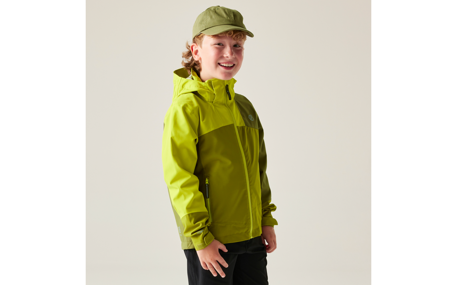 Kind in Dreiviertelansicht, trägt zweifarbige grüne Regenjacke mit Kapuze und grüne Kappe, schwarze Hose, heller Hintergrund