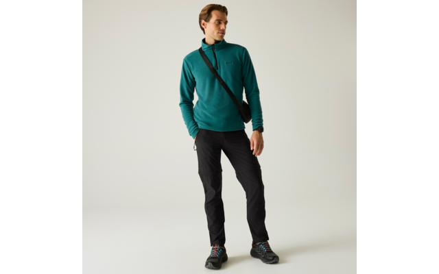 Person steht vor hellem Hintergrund, trägt türkisfarbenen Pullover, schwarze Hose, schwarze Umhängetasche und Wanderschuhe, blickt nach links