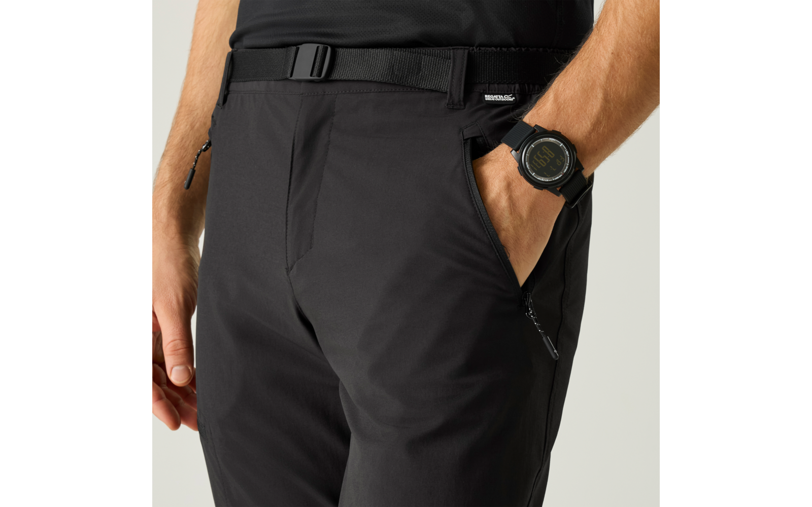 Hüft- und Oberschenkelbereich einer Person in schwarzen Hosen mit Gürtel; eine Hand in der Hosentasche, am Handgelenk eine schwarze Armbanduhr sichtbar, die andere Hand liegt locker am Körper