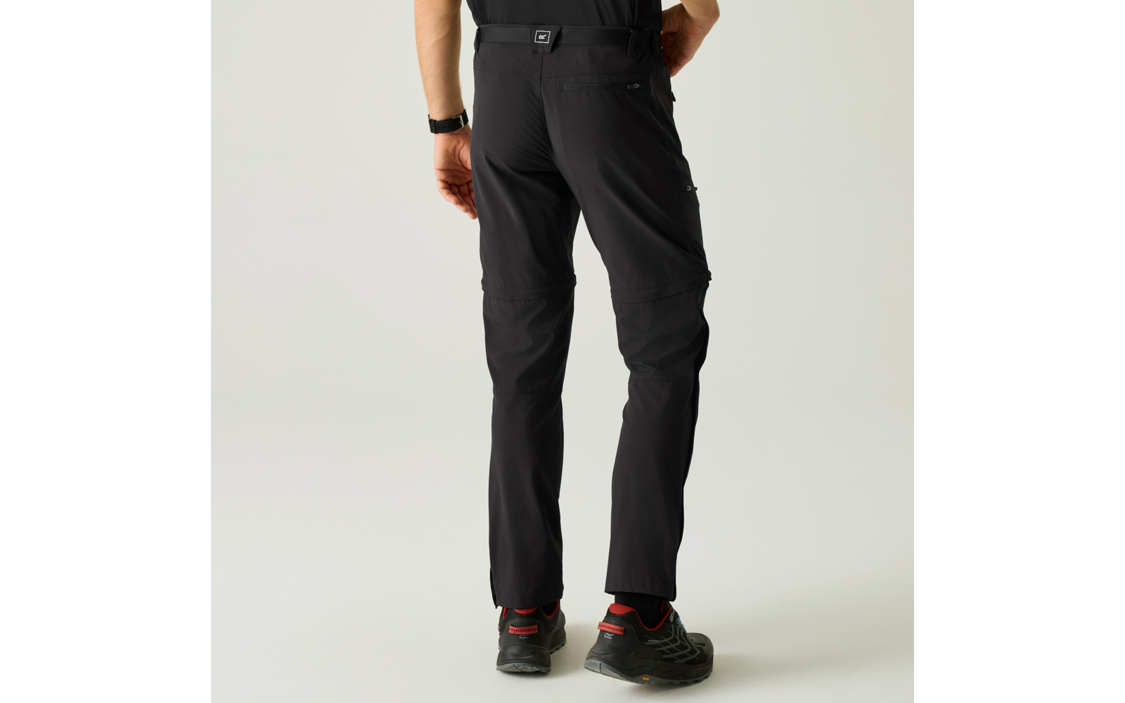 Person von hinten in schwarzen langen Hosen mit Naht am Knie, schwarzem Gürtel und schwarzen Sportschuhen mit roten Details vor hellem Hintergrund