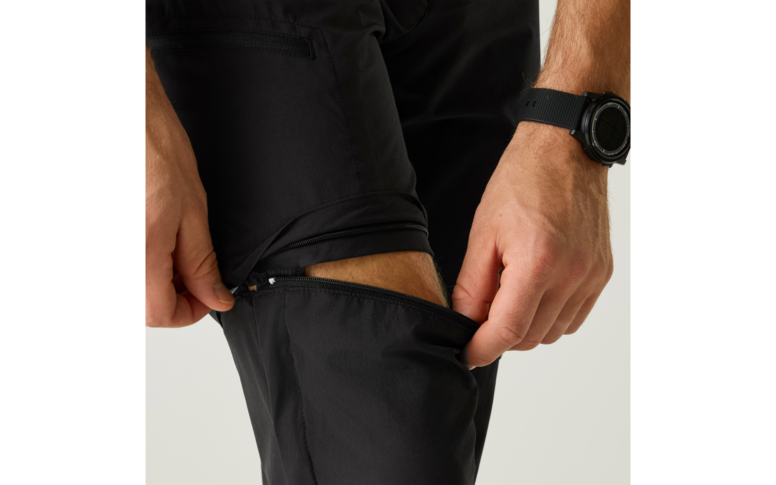 Hände an seitlichem Reißverschluss einer schwarzen Hose am Oberschenkel; Uhr am Handgelenk; heller Hintergrund