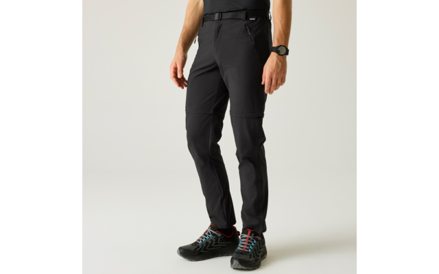 Untere Körperhälfte und Beine einer Person in schwarzen Hosen mit Reißverschluss am Oberschenkel, schwarzem Gürtel, Uhr am Handgelenk und schwarzen Schuhen mit roten Schnürsenkeln