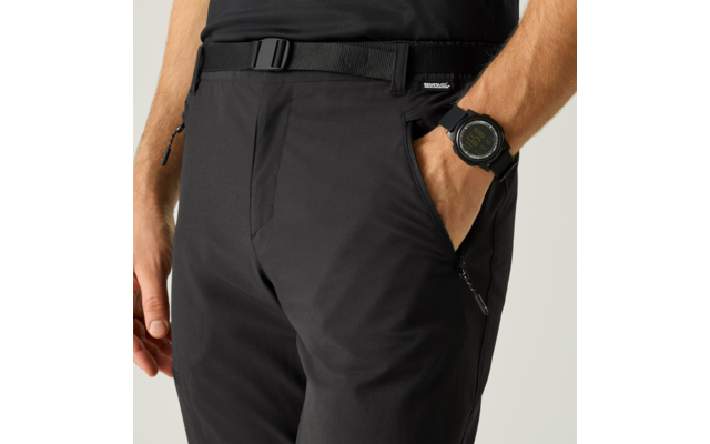 Hüft- und Oberschenkelbereich einer Person in schwarzen Hosen mit Gürtel; eine Hand in der Hosentasche, am Handgelenk eine schwarze Armbanduhr sichtbar, die andere Hand liegt locker am Körper