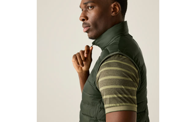 Person im Profil, trägt gesteppte grüne Weste und kurzärmliges gestreiftes grünes T-Shirt, Hand an Reißverschluss, heller einfarbiger Hintergrund