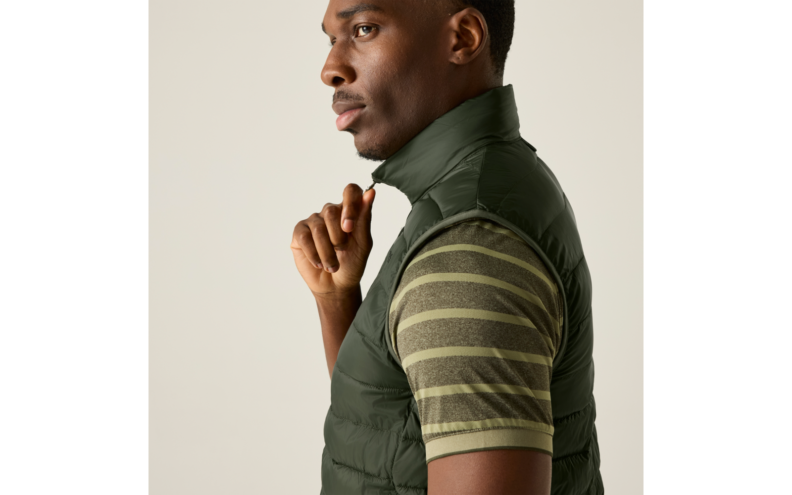 Person im Profil, trägt gesteppte grüne Weste und kurzärmliges gestreiftes grünes T-Shirt, Hand an Reißverschluss, heller einfarbiger Hintergrund