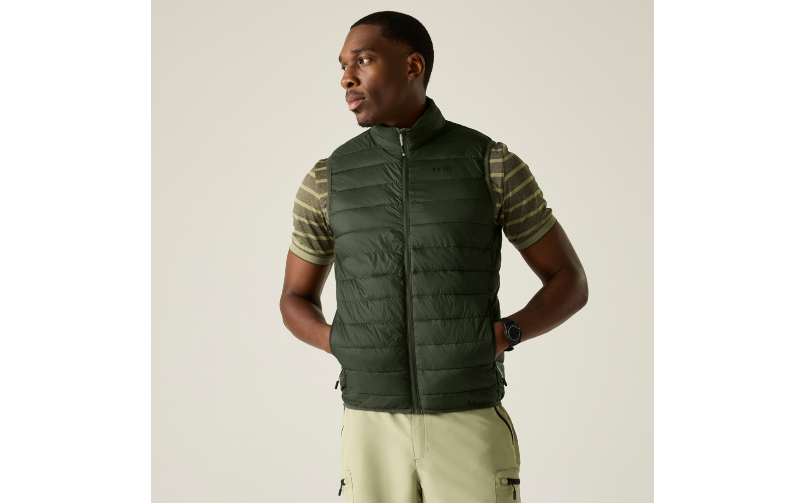 Mann trägt dunkelgrüne gesteppte Weste über gestreiftem T‑Shirt mit kurzen Ärmeln, helle Hose, Hände in den Taschen, Blick nach links vor einfarbig hellem Hintergrund
