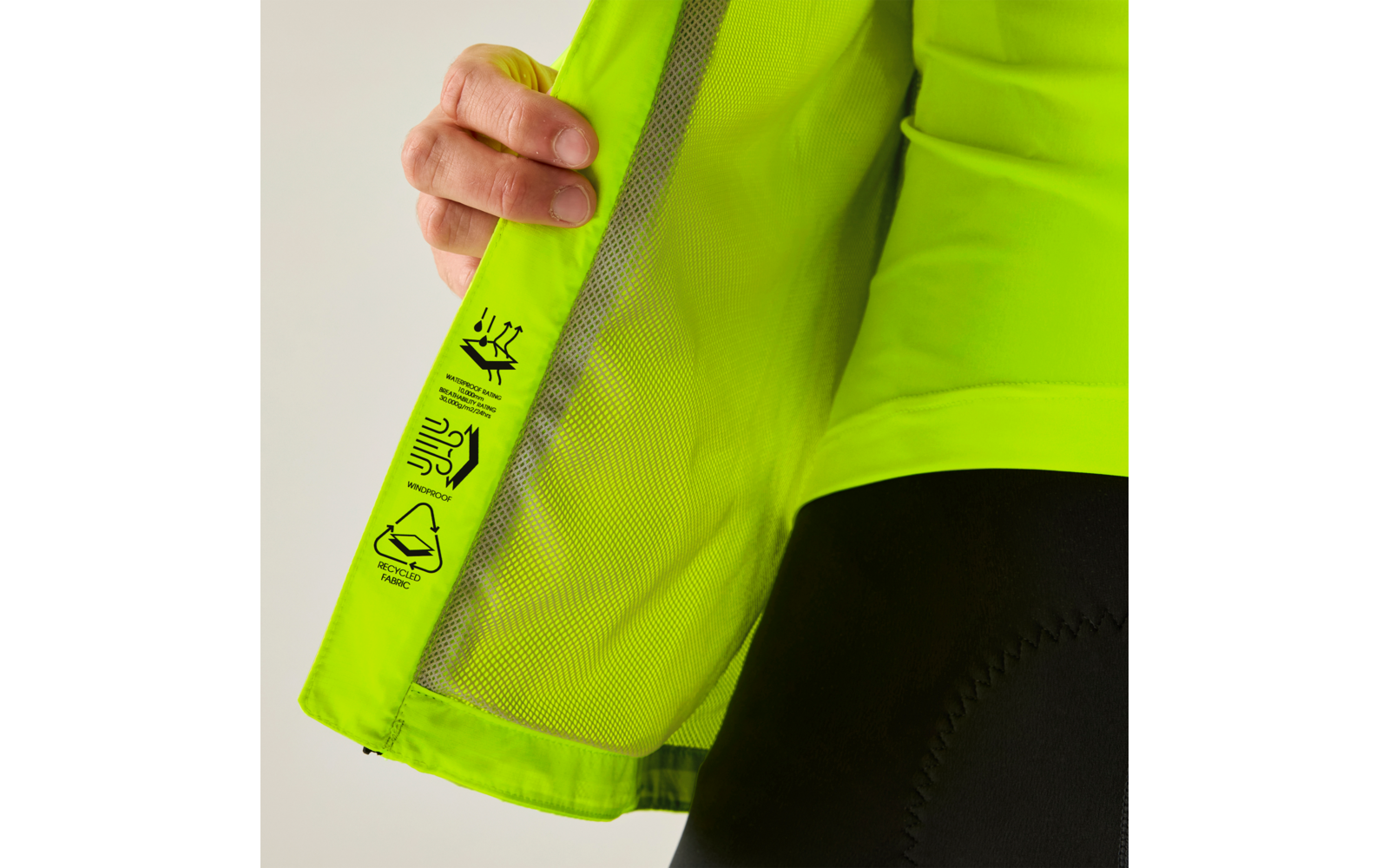 Hand hält eine aufgeklappte neongrüne Jacke, sichtbares Innenfutter aus Mesh, Pflegesymbole am Saum, Teil einer schwarzen enganliegenden Hose