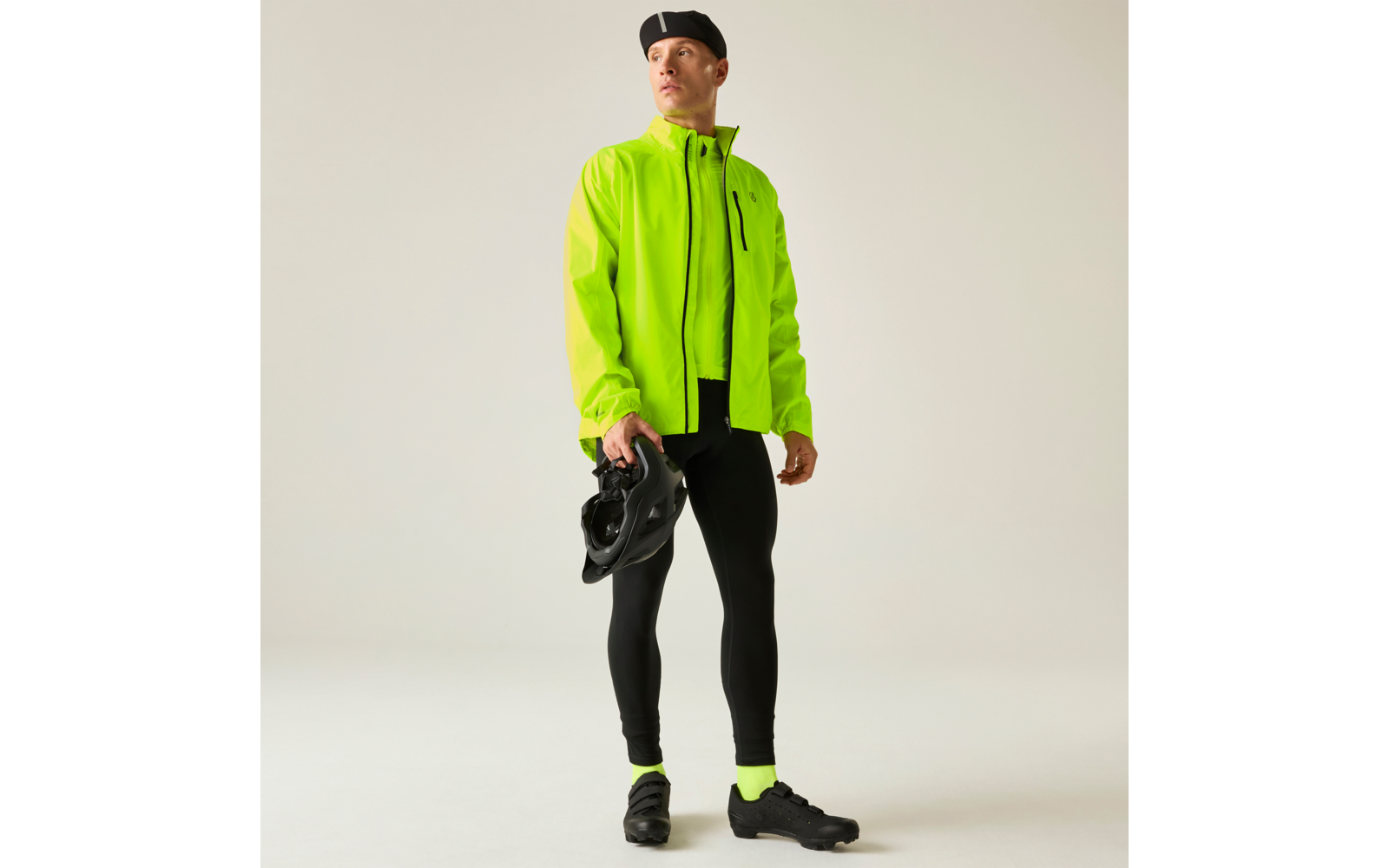 Person in neongelber Jacke, schwarzer Radbekleidung und schwarzem Barett, hält einen schwarzen Fahrradhelm und steht vor hellem Hintergrund