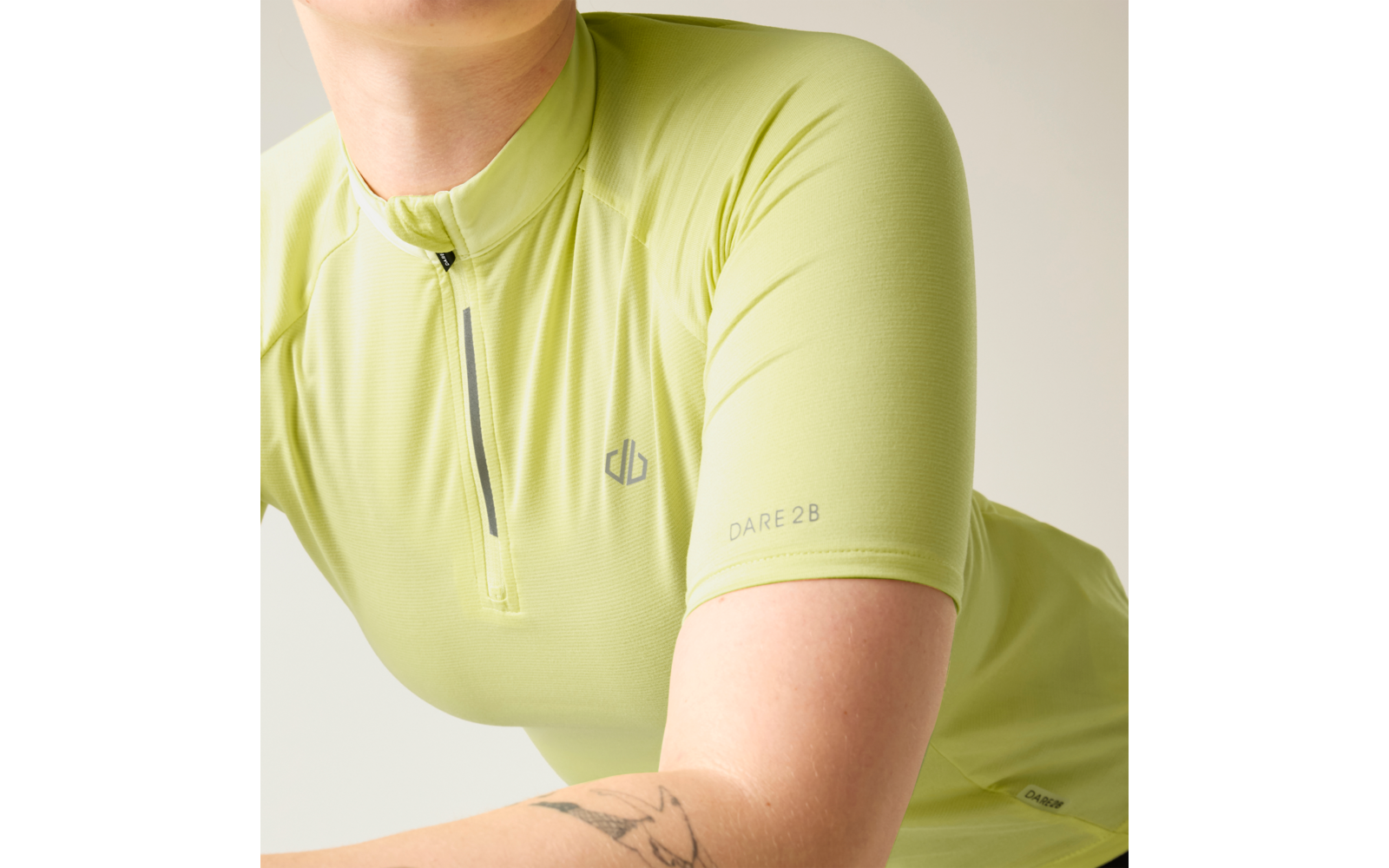 Hellgrünes kurzärmeliges Oberteil mit halbem Reißverschluss, kleinem Brustlogo und 'DARE 2 B' auf dem Ärmel; tätowierter Unterarm; heller einfarbiger Hintergrund
