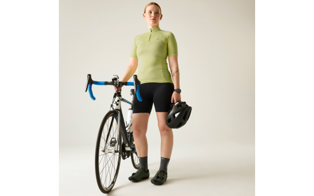 Person steht neben einem Rennrad und hält einen schwarzen Fahrradhelm, trägt hellgrünes kurzärmeliges Trikot, schwarze Radhosen und grüne Schuhe, sichtbare Tätowierung am Unterarm, Rennrad mit blau umwickeltem Lenker vor hellem neutralem Hintergrund