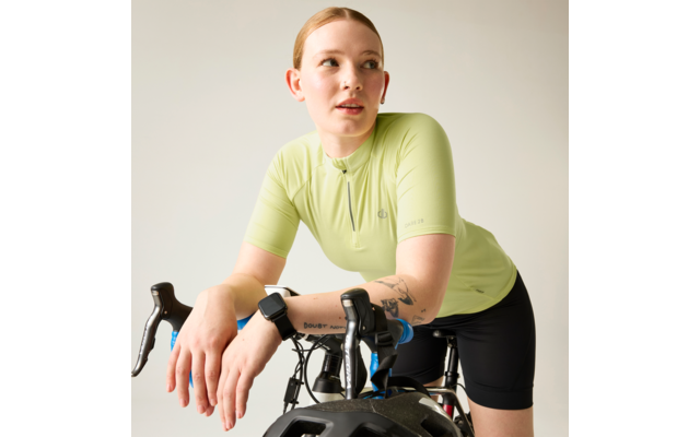 Person lehnt sich an den Lenker eines Rennrads, trägt hellgrünes kurzärmeliges Trikot mit Reißverschluss, schwarze Radhose, Smartwatch am linken Handgelenk, Tätowierung am Unterarm, schwarzer Fahrradhelm am Lenker, heller einfarbiger Hintergrund