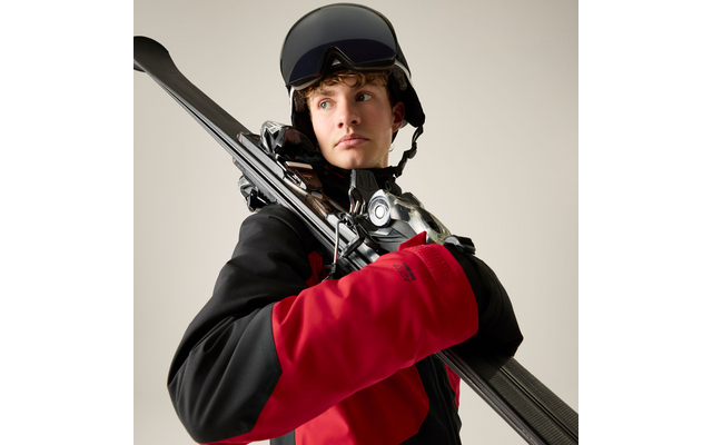 Person mit Skihelm und Skibrille, rot-schwarze Skijacke, trägt Skier über der Schulter und blickt seitlich