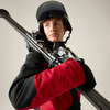 Person mit Skihelm und Skibrille, rot-schwarze Skijacke, trägt Skier über der Schulter und blickt seitlich