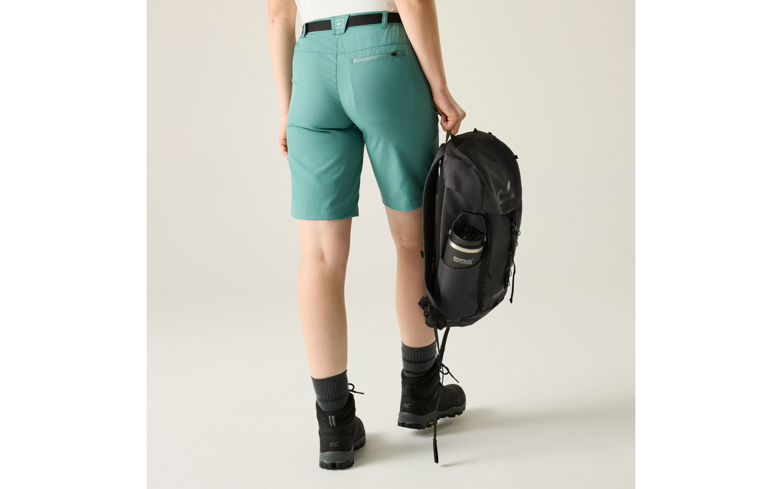 Rückansicht einer Person mit türkisgrünen Shorts, weißem Gürtel, dunklen Wanderschuhen und Socken, die einen schwarzen Rucksack in der rechten Hand hält