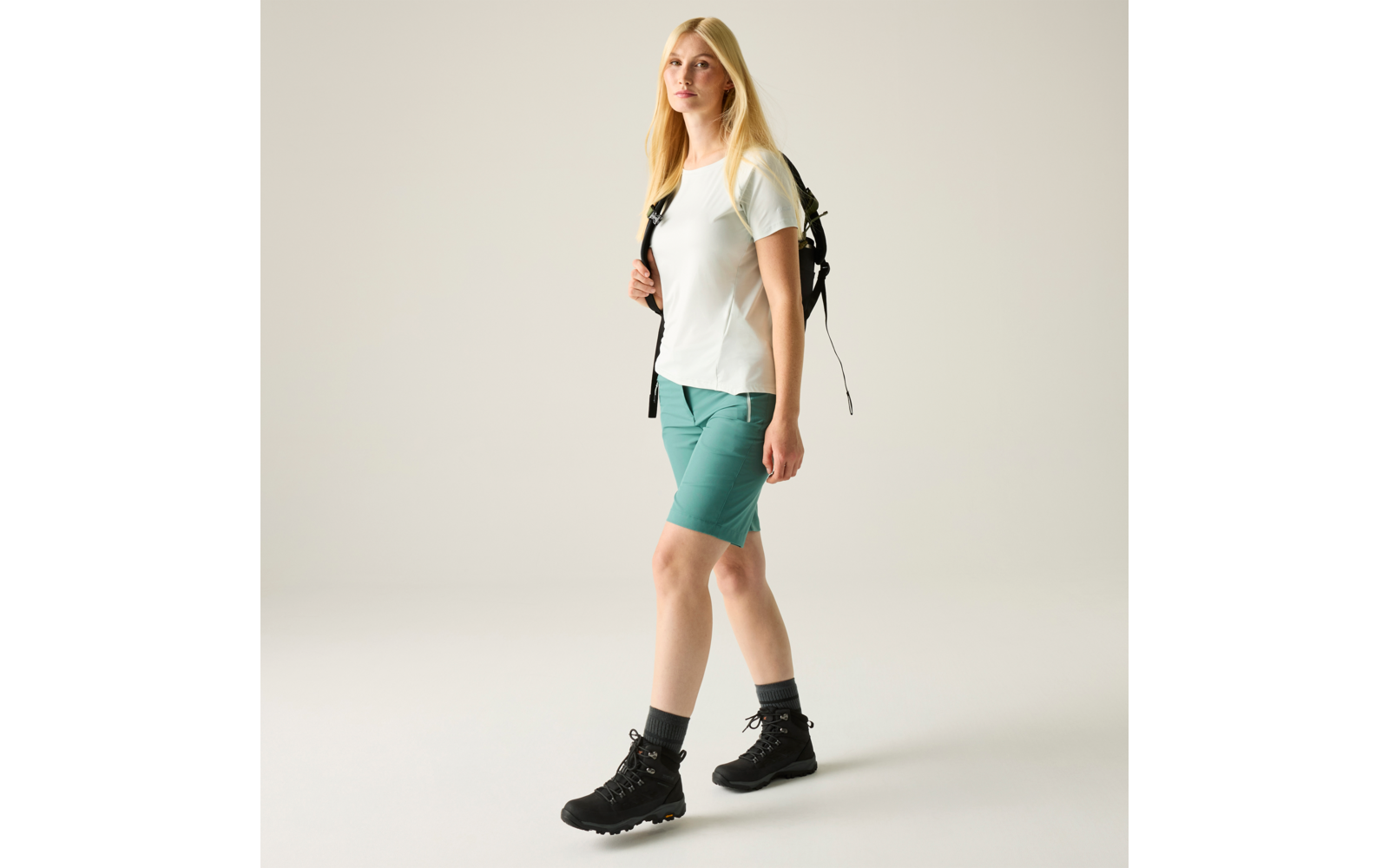 Person mit blondem Haar, weißem T-Shirt, türkisfarbenen Shorts, schwarzem Rucksack und schwarzen Wanderschuhen, stehend vor hellem Hintergrund
