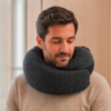 Homme aux cheveux foncés et courte barbe, coussin de voyage gris foncé autour du cou, pull beige, regard vers le bas, fond en panneaux de bois