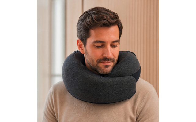 Homme aux cheveux foncés et courte barbe, coussin de voyage gris foncé autour du cou, pull beige, regard vers le bas, fond en panneaux de bois