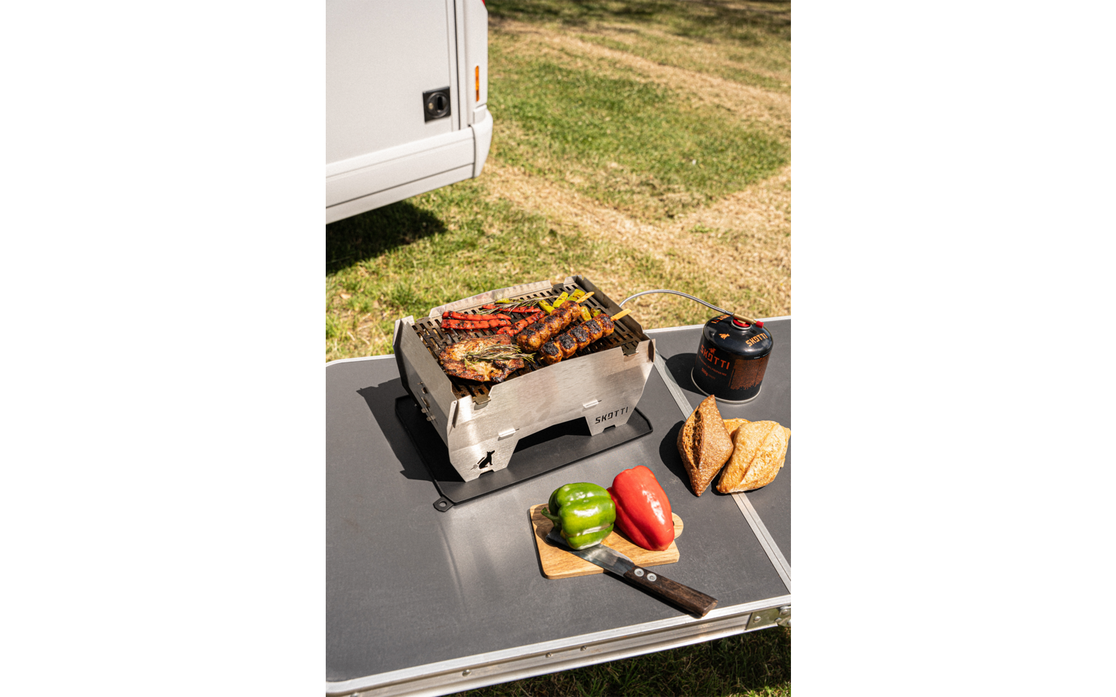 Ein Grill mit Gemüse auf einem Tisch im Freien, daneben eine Hand mit einem Grillbesteck