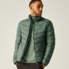Person in grüner gesteppter Jacke und grünem Pullover, Hände in den Taschen, Blick nach links vor hellem Hintergrund