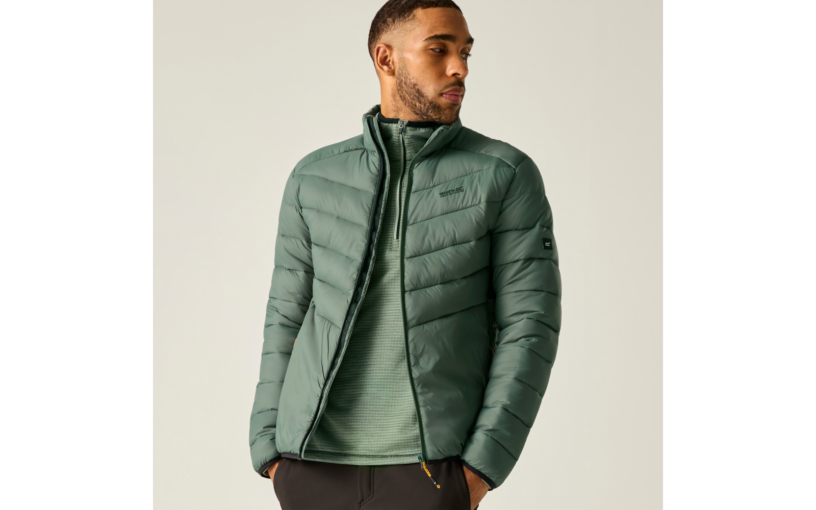 Person in grüner gesteppter Jacke und grünem Pullover, Hände in den Taschen, Blick nach links vor hellem Hintergrund