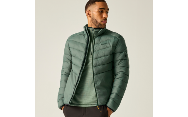 Person in grüner gesteppter Jacke und grünem Pullover, Hände in den Taschen, Blick nach links vor hellem Hintergrund