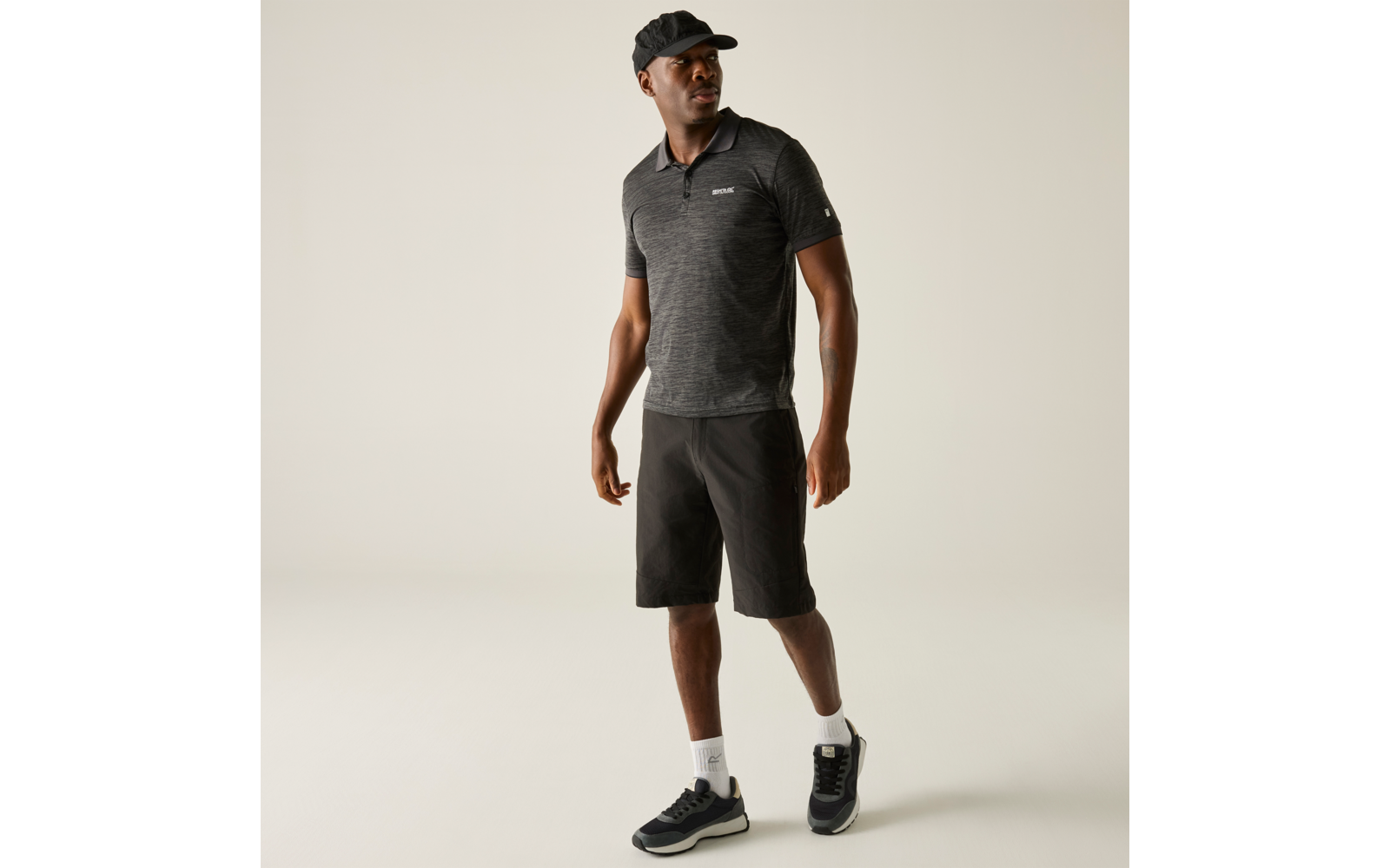 Persoon staand, licht gedraaid voor een lichte achtergrond, draagt een donker poloshirt, donkere knielange shorts, een zwarte pet, witte sokken en zwarte sneakers, kijkt opzij